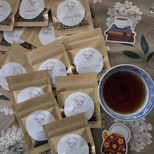 Puede incluir: Una variedad de productos de té, que incluyen pequeñas bolsas de papel marrón con etiquetas blancas y varias mezclas de té. También hay una taza de té con té y pegatinas decorativas de una taza y una tetera. Las etiquetas incluyen texto como "Immune Tea" y "Lady Grey".
