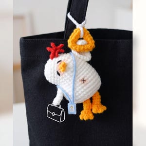 Pode incluir: Um porta-chaves de galinha em croché branco com crista vermelha, bico laranja e pés laranja. A galinha usa um cordão azul claro com uma pequena etiqueta e segura uma pasta bordada branca. Está pendurado num saco preto por um anel laranja.