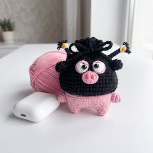 May include: A small, handmade crochet pouch featuring a black top with a drawstring closure and a pink bottom. The pouch has a character face with large white eyes, a pink snout, and two small black antennae topped with tiny white and yellow flowers.