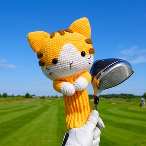 Pode incluir: Uma capa de crochê para taco de golfe em forma de gato malhado amarelo e branco com listras castanhas, olhos pretos e nariz bordado. A capa apresenta um corpo amarelo canelado e patas brancas, concebida para um driver de golfe de metal.