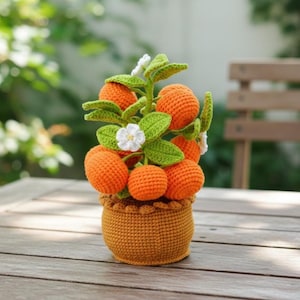 May include: A crocheted orange tree with bright orange fruit, green leaves, and white flowers, all contained in a brown crocheted pot. The handmade tree is displayed on a wooden table, with a blurred green background.