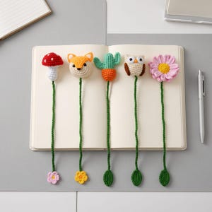 Op de afbeelding: Vijf handgemaakte gehaakte boekenleggers met een rood-witte paddenstoel, een oranje-crèmekleurige vossenkop, een groene cactus in een oranje pot, een bruin-crèmekleurige uil en een roze bloem, elk met een lange groene steel en een kleine voet.