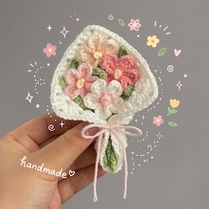 May include: A small crochet flower bouquet featuring white, peach, and pink yarn flowers with green leaves, wrapped in a white crocheted square base tied with a thin pink string. The word handmade is written in white cursive text on the left side.