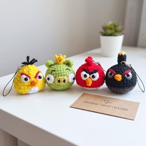 Op de afbeelding: Vier gehaakte pluche sleutelhangers met ronde lichamen, expressieve ogen en kleine snavels. De set bevat een gele vogel, een groen varken met een kroon, een rode vogel en een zwarte vogel. Een bruine kaart bevat de tekst HANDMADE CROCHET CREATIONS.