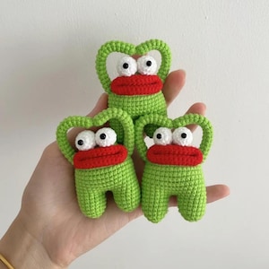 May include: Three small, handmade crochet figures in a bright lime green yarn. Each figure features a large, thick red mouth, two prominent white eyes with black pupils, and a rounded, arched top section that forms a handle-like shape.