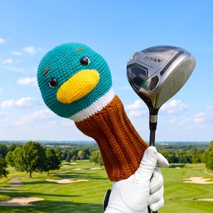 May include: A crochet golf club head cover shaped like a mallard duck head with a teal top, yellow beak, and brown neck, positioned next to a silver and black Titan golf driver club head against a clear blue sky and a green golf course background.