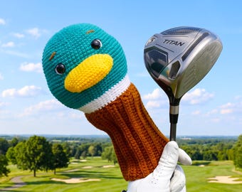 May include: A crochet golf club head cover shaped like a mallard duck head with a teal top, yellow beak, and brown neck, positioned next to a silver and black Titan golf driver club head against a clear blue sky and a green golf course background.