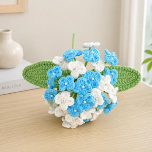 Pode incluir: Um buquê de hortênsias de crochê feito à mão com flores azuis e brancas com detalhes de pérolas. As flores são dispostas em uma base de folha verde. Um arranjo floral decorativo para casa.