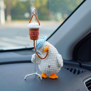 May include: A white crocheted duck figurine wearing a light blue hat and orange feet, standing next to a silver metal stand. A small crocheted coffee cup in white and brown hangs from the stand, connected to the duck by a brown yarn tube.