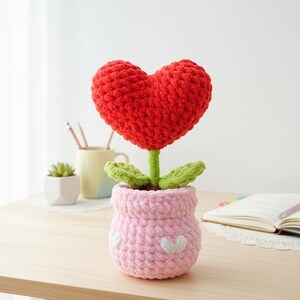 May include: A crocheted flower with a red heart-shaped bloom, green leaves, and a pink pot. The pot is decorated with white heart-shaped accents. The flower sits on a light-coloured wooden surface.