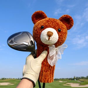 May include: A brown crocheted bear golf club headcover featuring a white muzzle, black eyes, and a sheer white ribbon bow. The bear headcover is positioned next to a dark grey golf club head, with the texture of the yarn clearly visible.