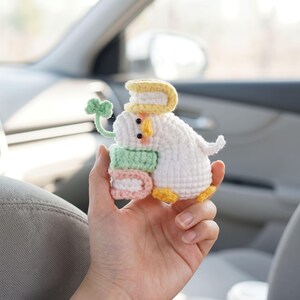 May include: A small white crocheted duck figurine featuring a yellow beak and orange feet. The duck holds a stack of two small books in light green and pink, has a yellow book on its head, and a green four-leaf clover stem extending from its side.
