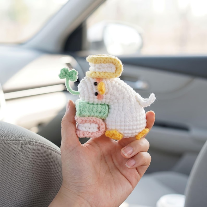 May include: A small white crocheted duck figurine featuring a yellow beak and orange feet. The duck holds a stack of two small books in light green and pink, has a yellow book on its head, and a green four-leaf clover stem extending from its side.