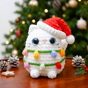 May include: A white crocheted cat plush toy wearing a red Santa hat with a white pom-pom. The cat features large dark blue eyes, a small pink nose, and is wrapped in a green yarn string decorated with small, colorful crocheted light bulbs.