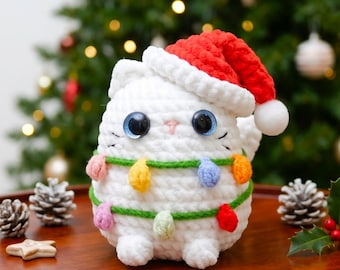 May include: A white crocheted cat plush toy wearing a red Santa hat with a white pom-pom. The cat features large dark blue eyes, a small pink nose, and is wrapped in a green yarn string decorated with small, colorful crocheted light bulbs.