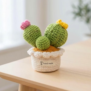 Könnte beinhalten: Eine gehäkelte Kaktuspflanze mit drei grünen Segmenten, einer rosa Blüte links und einer gelben Blüte rechts. Der Kaktus steht in einem cremefarbenen gehäkelten Topf mit weißem, gewelltem Rand und einem Etikett mit der Aufschrift hand made.