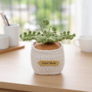 May include: A handmade crochet plant in a white pot. The plant has green leaves and a brown base. The pot has a small tag that reads "Hand Made". The plant is sitting on a wooden surface.