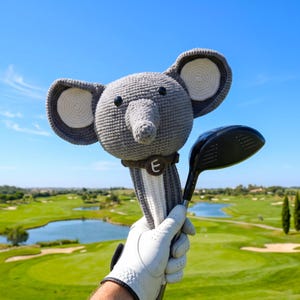 May include: A gray and white crocheted elephant golf club headcover featuring large ears, black bead eyes, and a dark brown collar with a white letter E. The headcover is attached to a black golf club driver with a long, slender metal shaft.