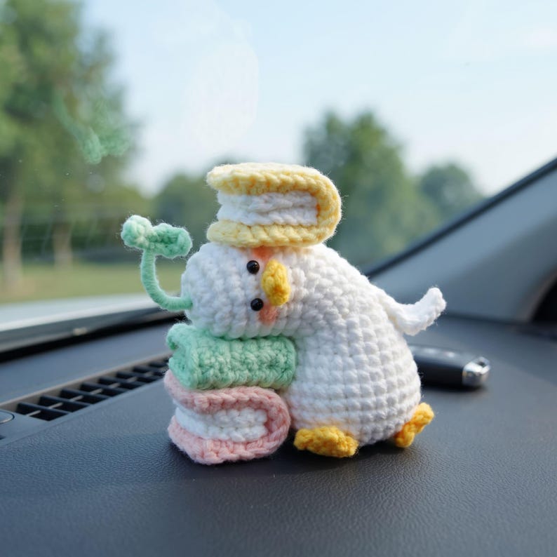 May include: A white crocheted duck figurine with a yellow beak and feet, featuring a small green sprout on its head. The duck is holding a stack of two books in light green and pink, with a yellow book balanced on top of its head.