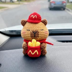 Op de afbeelding: Een gehaakte capybara knuffel met een rode pet met een geel M-logo en een rode sjaal. De capybara houdt een rode houder met een geel M-logo vast, gevuld met gele gehaakte frietjes. Het speelgoed heeft bruine vacht en gesloten ogen.