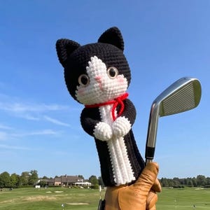 May include: A black and white crocheted cat golf club headcover featuring large dark eyes, a pink nose, and a red cord tied around its neck. The cat has pointed ears and white paws, and it is positioned next to a silver metal golf club iron.