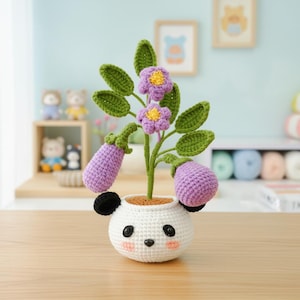 May include: A handmade crochet plant with purple flowers, green leaves, and two purple eggplant-shaped fruits. The plant is in a white panda-shaped pot with black ears and a smiling face. The plant is on a wooden surface.
