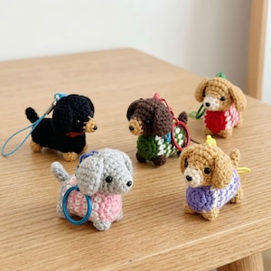 Puede incluir: Cinco llaveros de perro salchicha de ganchillo en varios colores. Los perros tienen suéteres de diferentes colores, incluyendo rosa, rojo, verde y morado. Cada perro tiene un llavero o correa.