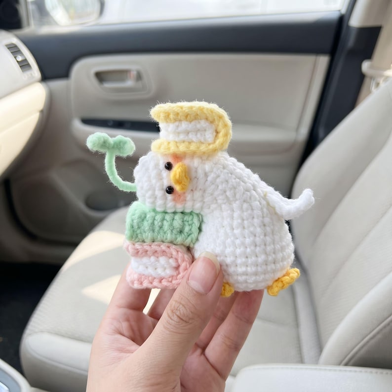 May include: A white crocheted duck figurine featuring a yellow beak and feet, black eyes, and pink cheeks. The duck holds a stack of two small books in light green and pink, with a yellow book on its head and a small green sprout extending outward.
