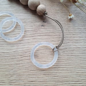 Könnte beinhalten: Nahaufnahme eines Schnullerclips mit drei durchscheinenden Ringen und Holzperlen. Der Clip hat eine neutrale Farbpalette und befindet sich auf einer hellen Holzmaserungsoberfläche. Getrocknete Blumen sind im Hintergrund.