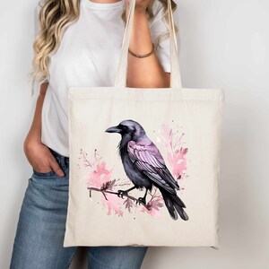 May include: A tote bag with a black crow with pink watercolor floral accents. The crow is perched on a branch with pink flowers and leaves.