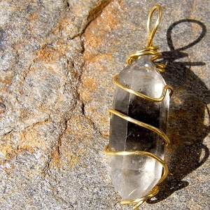May include: A clear quartz crystal wrapped in gold wire, creating a pendant. The pendant is photographed on a rough, grey and brown stone surface.