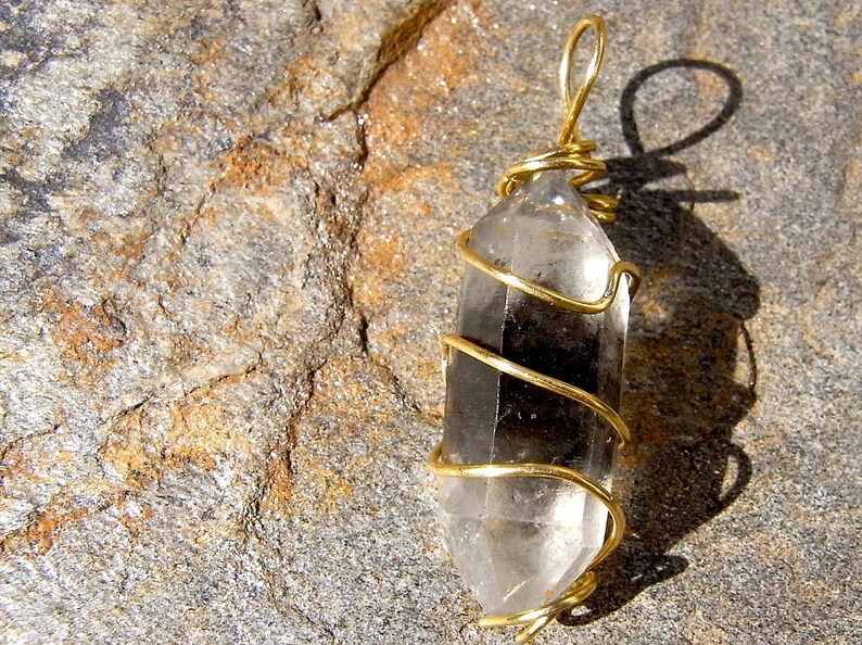 May include: A clear quartz crystal wrapped in gold wire, creating a pendant. The pendant is photographed on a rough, grey and brown stone surface.