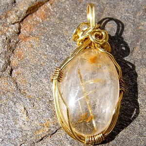 May include: Gold wire wrapped pendant featuring a clear quartz crystal with gold inclusions. The crystal is oval shaped and has a wire bail for hanging.