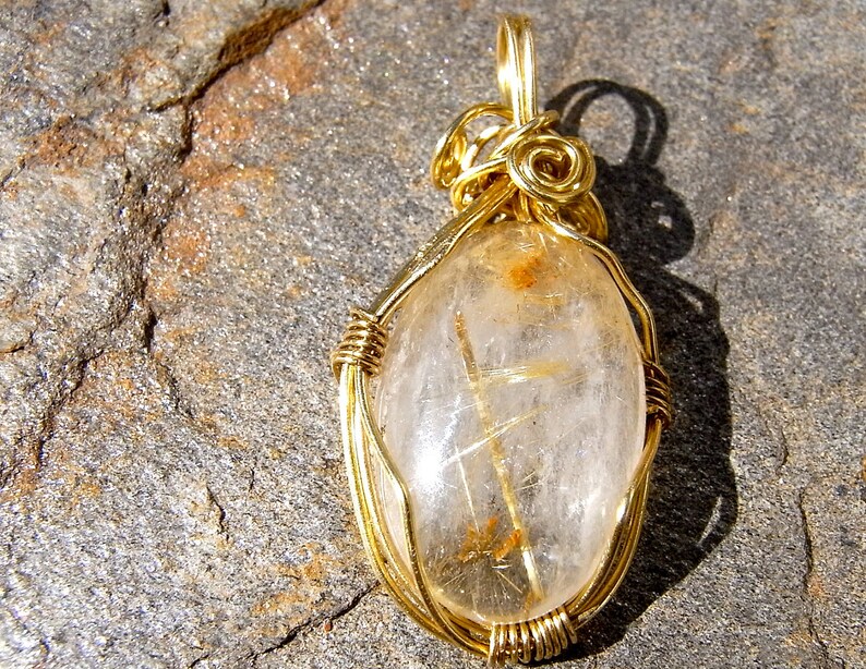 May include: Gold wire wrapped pendant featuring a clear quartz crystal with gold inclusions. The crystal is oval shaped and has a wire bail for hanging.
