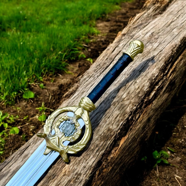 May include: A decorative sword with a gold-colored hilt and accents, a black handle, and a silver-colored blade. The sword is resting on a weathered wooden log, with green grass in the background.