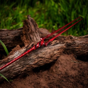 May include: A red spear with a spiraled handle and two long, pointed tines. The spear is made of metal and is set against a backdrop of weathered wood and green foliage.