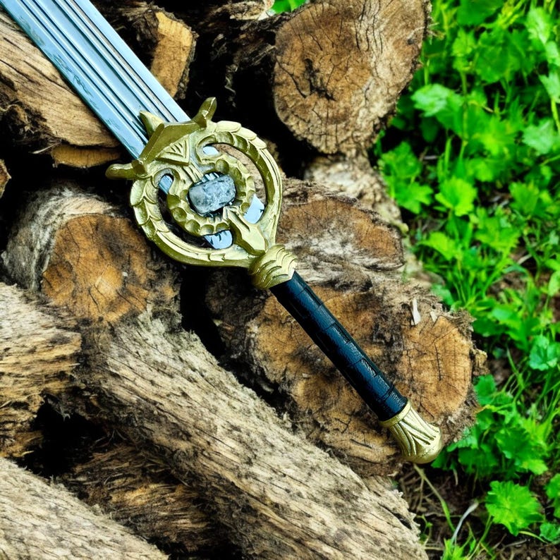May include: A decorative sword with a silver blade and a gold-colored hilt. The hilt features an ornate design with a black handle. The sword is resting on a pile of wooden logs, with green foliage in the background.