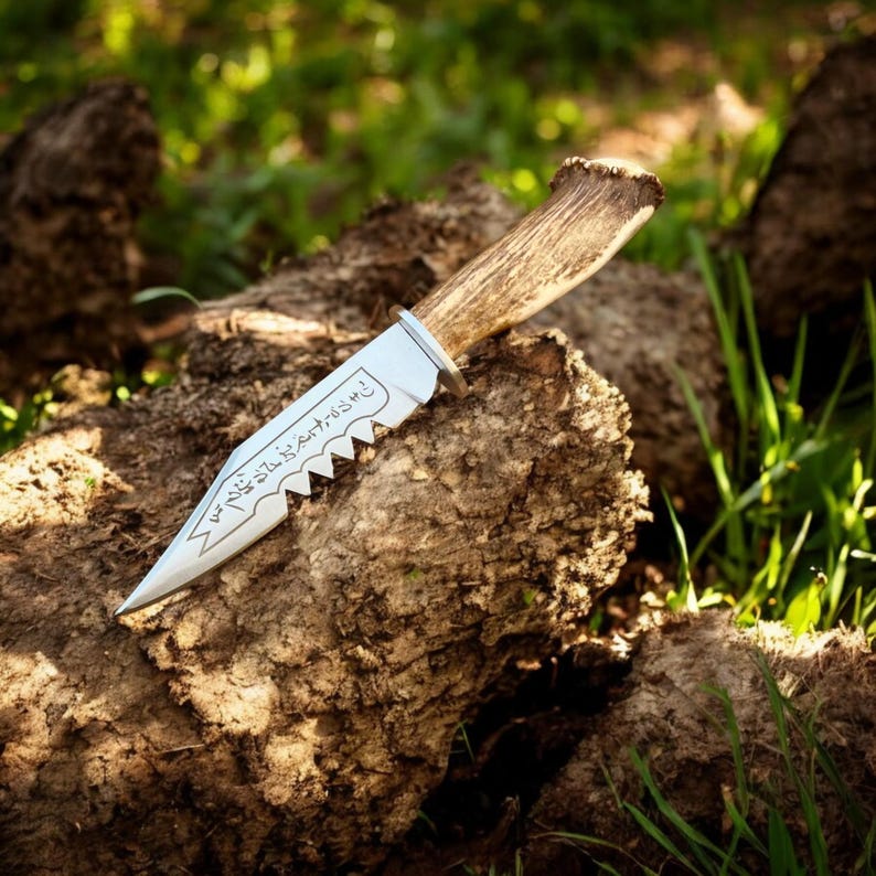 May include: A hunting knife with a silver blade and a wooden handle. The blade has a serrated edge and etched text. The handle is made of antler. The knife rests on a piece of wood, with green grass in the background.