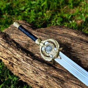 May include: A decorative sword with a silver blade and a gold-colored hilt. The hilt features an ornate design with a central stone. The handle is wrapped in black material. The sword is displayed on a weathered wooden surface.
