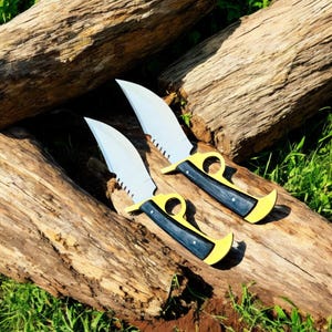 May include: Two knives with serrated blades and yellow handles with black grips rest on a weathered wooden surface. The blades are silver, and the handles have a finger loop design. The knives are displayed outdoors.