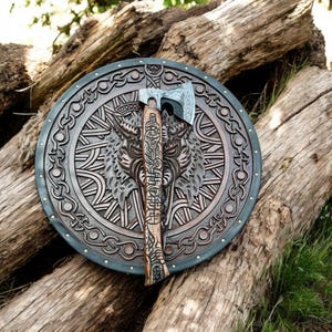 May include: A detailed, round shield with a wolf carving and an axe. The shield is brown with a dark blue border and brass studs. The axe has a wooden handle with carvings and a silver blade. The items are displayed on a pile of logs.