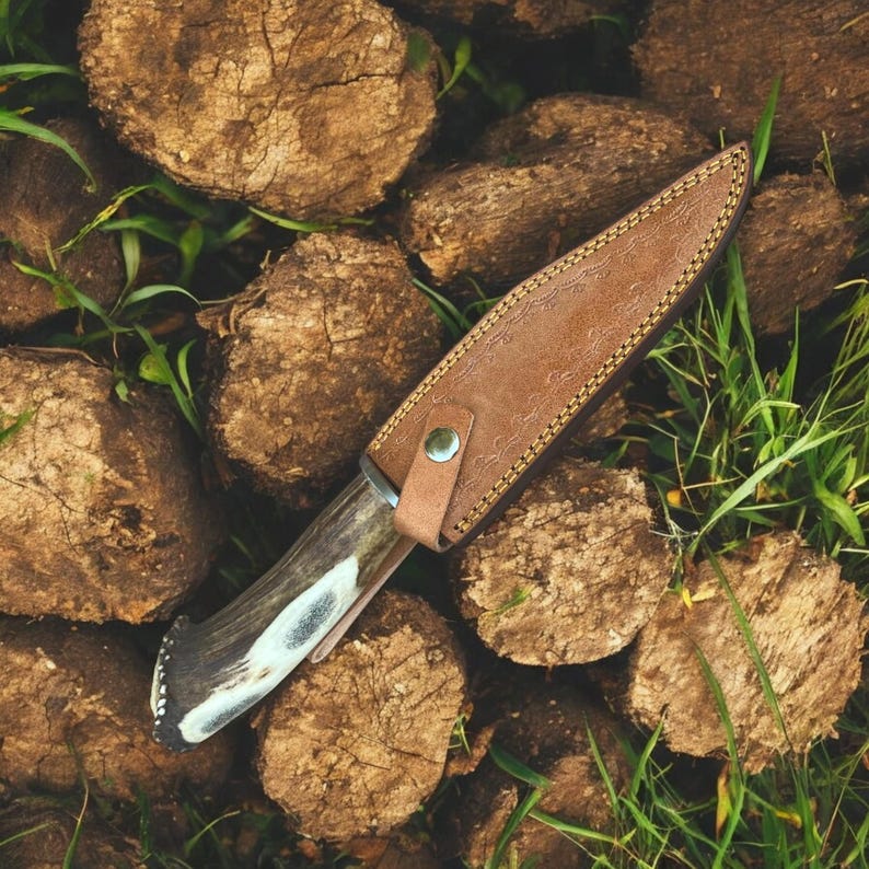 May include: A hunting knife with a brown leather sheath. The knife has a dark handle with white accents and a silver blade. The sheath is tan with decorative stitching and a snap closure. The knife rests on a bed of wood and grass.