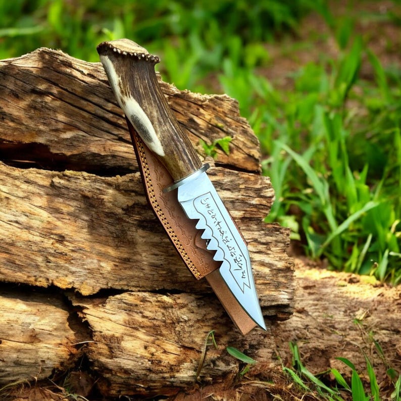 May include: A hunting knife with a serrated blade and a handle made from antler material. The knife is in a brown leather sheath and rests on a piece of wood. The blade has etched text.