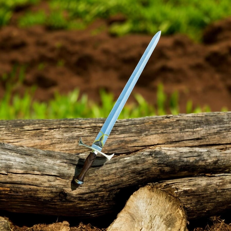 May include: A long, silver sword with a light blue blade and a dark brown wooden handle. The hilt is silver with a decorative design. The sword is resting on a large, weathered log, with a blurred green and brown background.