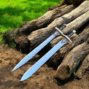 May include: Two swords with light blue blades and gold-colored crossguards, each featuring a crest. The hilts are black with gold accents. The swords are resting on weathered wooden logs, with green grass in the background.