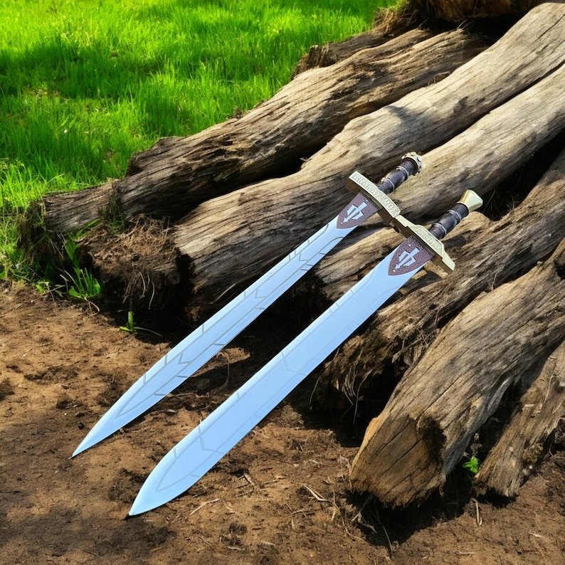 May include: Two swords with light blue blades and gold-colored crossguards, each featuring a crest. The hilts are black with gold accents. The swords are resting on weathered wooden logs, with green grass in the background.