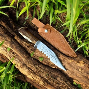May include: A hunting knife with a wooden handle and a silver blade, resting on a piece of wood. The knife has a serrated edge and is accompanied by a brown leather sheath. The blade has etched text.