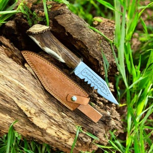 May include: A hunting knife with a serrated blade and a handle made of antler. The knife is displayed with a brown leather sheath. The blade has etched text. The knife and sheath are resting on a piece of wood.
