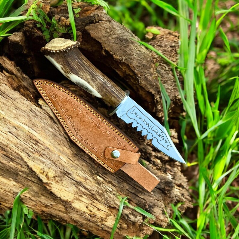 May include: A hunting knife with a serrated blade and a handle made of antler. The knife is displayed with a brown leather sheath. The blade has etched text. The knife and sheath are resting on a piece of wood.