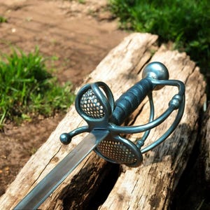 Könnte beinhalten: Ein Rapier-Schwert aus Stahl mit einem komplexen Parier und einem lederumwickelten Griff. Die Klinge ist gerade und scharf, und der Parier weist ein perforiertes Korbmuster auf. Das Schwert liegt auf einer verwitterten Holzoberfläche, mit grünem Gras im Hintergrund.